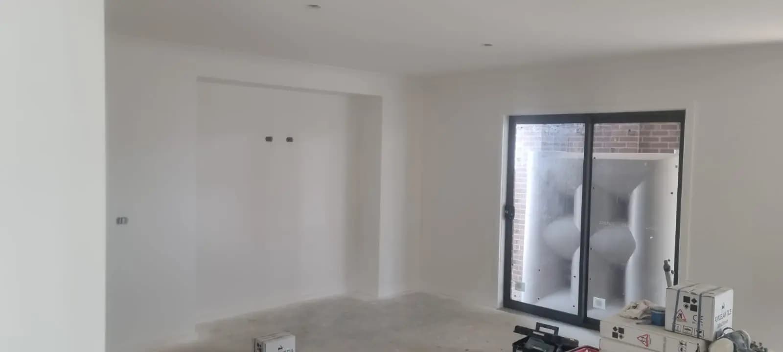 Living area of a new house with walls and ceiling painted white, a recessed alcove in the far wall and a black-framed sliding door looking onto a brick courtyard.