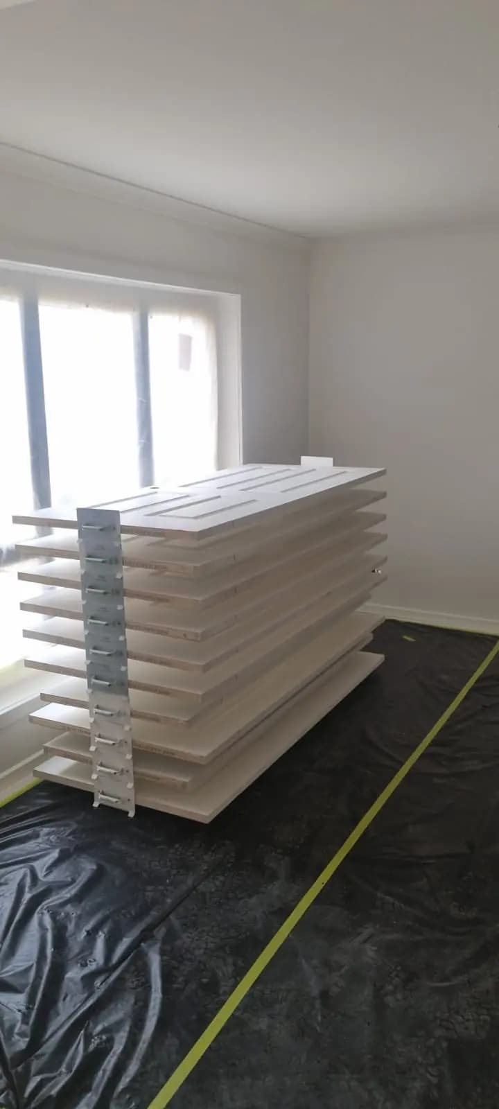 A stack of freshly painted white doors and shelves on a drying rack in an empty room, standing on a black drop sheet taped at the edges.