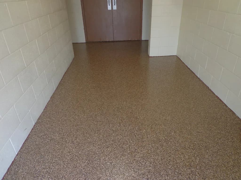 A tan and brown flake epoxy floor running down a hallway between painted block walls towards a pair of timber doors.